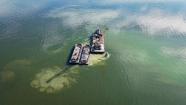 An aerial photo of a tug pushing a barge with a tall crane. Attached to the crane is a grapple that looks to be dropping material into the water, which is churned up. A barge loaded with cobble is next to the barge with the crane. On the other side of the cobble barge is an empty barge with an idle front-end loader.